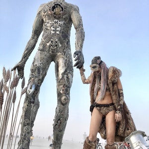 Puede incluir: Una gran escultura de metal de una figura humana se alza en un paisaje des&eacute;rtico. La escultura est&aacute; hecha de varias partes met&aacute;licas y tiene un aspecto desgastado e industrial. Una persona con un chaleco de piel y una m&aacute;scara se encuentra junto a la escultura, sosteniendo su mano.