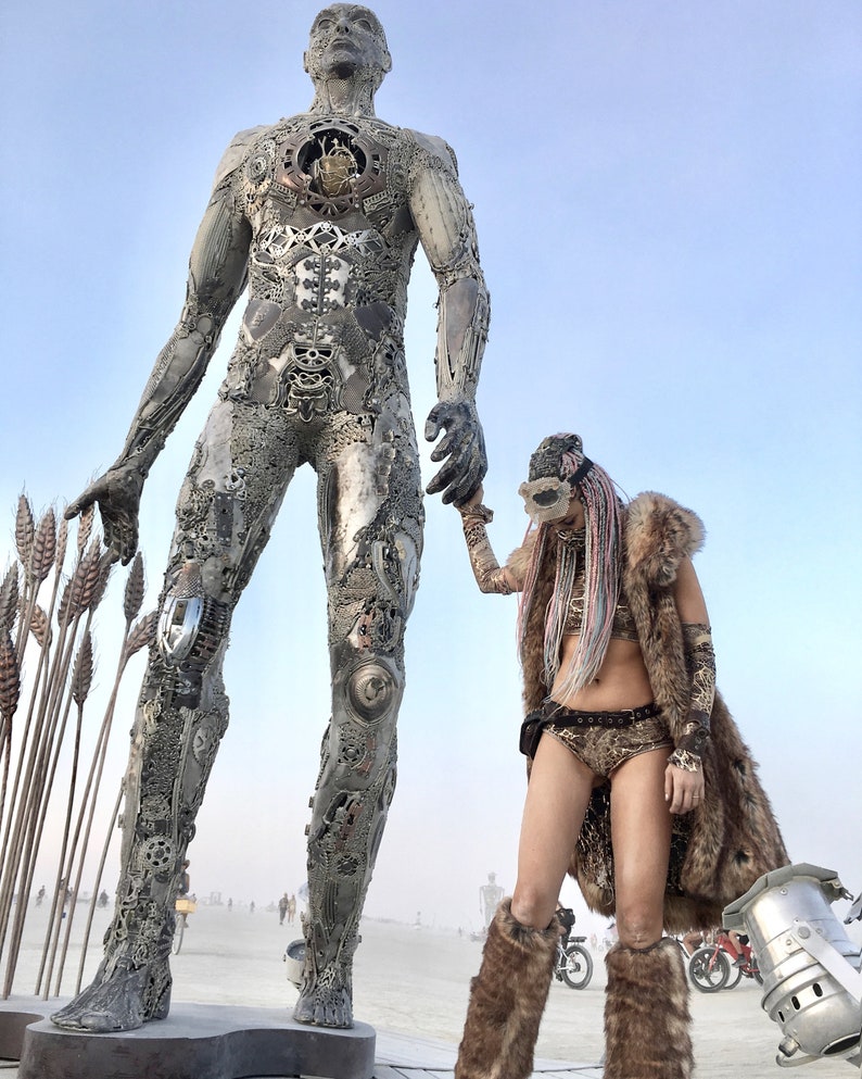 Puede incluir: Una gran escultura de metal de una figura humana se alza en un paisaje des&eacute;rtico. La escultura est&aacute; hecha de varias partes met&aacute;licas y tiene un aspecto desgastado e industrial. Una persona con un chaleco de piel y una m&aacute;scara se encuentra junto a la escultura, sosteniendo su mano.