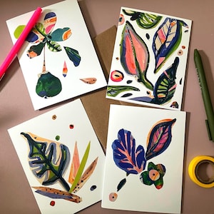 Puede incluir: Cuatro tarjetas de felicitación blancas con ilustraciones botánicas coloridas, que incluyen hojas y flores, están dispuestas sobre una superficie rosa. También hay un bolígrafo rosa, un bolígrafo verde y un rollo de cinta adhesiva amarilla. Las tarjetas presentan tonos vibrantes de azul, verde, rosa y naranja.