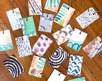 May include: A collection of 18 gift tags with various watercolor patterns. The tags are rectangular and have a hole punched in the top for string. The patterns include abstract designs, geometric shapes, and floral motifs. The colors are bright and vibrant, including blue, green, pink, and orange.