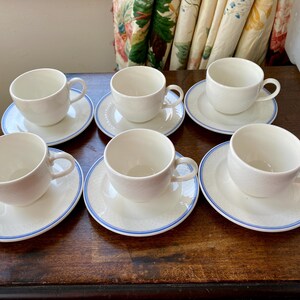 May include: Six white teacups with blue trim and matching saucers on a wooden surface. The teacups and saucers have a simple, elegant design.
