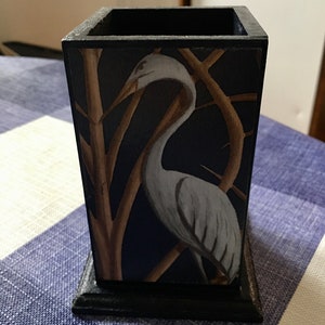 May include: A black wooden pencil holder featuring a blue and gold heron design. The holder has a square shape and a wide base.