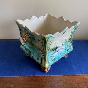 May include: A square ceramic planter with a scalloped rim. The planter has a relief design of swans, reeds, and water. The colours are cream, teal, and brown. The planter sits on small, decorative feet.