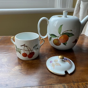 May include: A white porcelain tea set with gold detailing, including a teapot, a cup, and a lid. The set is decorated with illustrations of cherries and oranges. The teapot has a curved handle and spout. The cup has two handles.