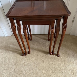 May include: A set of three dark wood nesting tables. Each table has a square top with a decorative edge. The tables are supported by curved legs with carved details. The tables are arranged in a nested configuration, a classic furniture set.