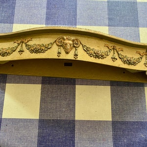 May include: A decorative wooden shelf with a curved top and ornate details. The shelf is painted in a light brown colour and features a gold-coloured ram's head design in the centre. The shelf is decorated with floral motifs and ribbons.
