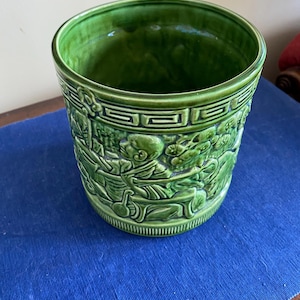 May include: A glossy green ceramic planter with a detailed relief design. The planter features a wide, open top and a cylindrical shape. The relief depicts figures and foliage. The planter is sitting on a blue surface.
