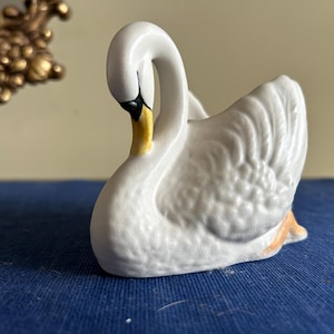 May include: A white ceramic swan figurine with a curved neck, a yellow and black beak, and textured wing details. The swan is depicted in a resting position with its head tucked slightly downward, showcasing a smooth, glossy finish throughout.