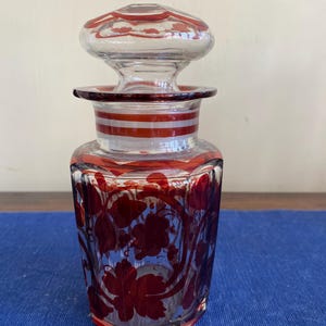 May include: A clear glass perfume bottle with a red floral design. The bottle has a matching stopper and red and white striped accents. The bottle's body is a faceted shape, and the floral design is a deep red colour.