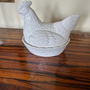May include: A white ceramic hen-shaped dish with a textured finish. The hen features a detailed comb, beak, and feather detailing, resting on a basket-style base. This decorative item is likely for serving or storage purposes.