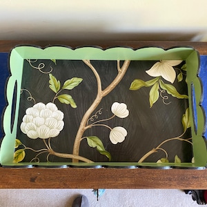 May include: Rectangular serving tray with a black background and a floral design. The tray has a scalloped edge with a green border. The floral design includes white flowers, green leaves, and brown branches.
