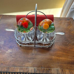 May include: A condiment server set featuring two clear glass bowls with fluted bases, each topped with a glass lid containing embedded orange and green fruit motifs. The bowls include matching glass spoons and sit in a chrome metal caddy.