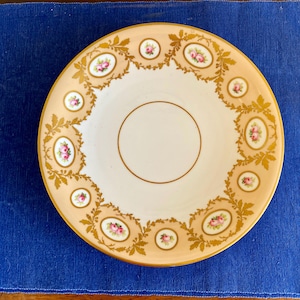 May include: A white porcelain saucer with a delicate floral design. The saucer features a gold rim and intricate floral patterns in shades of pink and gold.