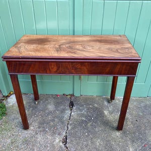May include: An antique wooden table with a rectangular top and dark brown finish. The tabletop displays a prominent wood grain pattern. The table has four slender legs and small caster wheels. The table is approximately 76 cm long.
