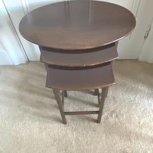 May include: A set of three nesting tables in a dark brown finish. The largest table has a round top, with the other two having a curved, rectangular shape. The tables are made of wood and have a classic design.