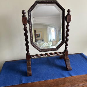 May include: A dark brown wooden swivel mirror featuring an octagonal frame with carved detailing. The mirror is supported by two vertical barley twist columns and a horizontal barley twist base, resting on two feet for stability on a flat surface.
