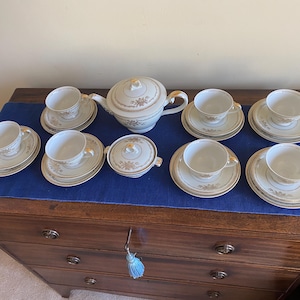 May include: A vintage tea set comprising a teapot, sugar bowl, and eight cups and saucers. The white porcelain has a gold trim and floral pattern. The set is arranged on a blue cloth on a wooden chest of drawers.