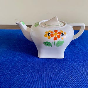 May include: White ceramic teapot with a square base, a curved spout, and a handle. The teapot is decorated with orange, yellow, and green floral designs. The lid has a small, triangular handle. The teapot is on a blue surface.