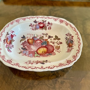 May include: Oval serving dish with a white background and a decorative fruit basket design. The rim is scalloped and features a claret floral pattern. The fruit basket includes apples, grapes, and peaches.