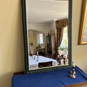 May include: Rectangular mirror with a dark green rope-effect frame and gold trim. The mirror reflects a dining room scene with a table set for a meal. The mirror is displayed on a wooden dresser covered with a blue cloth.