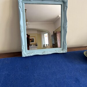 May include: A rectangular mirror with a pale blue ornate frame. The frame features decorative scrollwork at the corners and edges. The mirror reflects a room with a white ceiling and a window. The mirror is on a dark wooden surface.