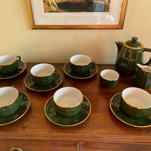 May include: A dark green tea set with gold edging, comprising cups, saucers, a teapot, a creamer, and a sugar bowl. The set is displayed on a wooden surface, with a framed picture in the background.