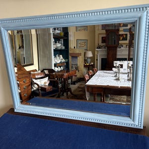 May include: A rectangular mirror with a pale blue frame. The frame has a beaded edge and a painted finish. The mirror reflects a room with furniture and decor.