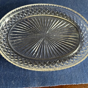 May include: An oval clear glass serving platter featuring a central sunburst pattern radiating from the middle, surrounded by a textured diamond crosshatch border along the rim. The edges of the platter have a scalloped, serrated design.