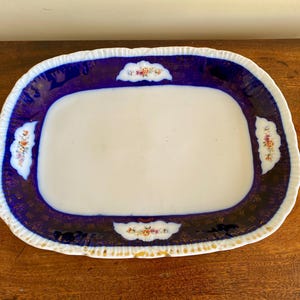 May include: A white oval serving platter with a blue and gold rim and floral designs. The platter is on a wooden surface.