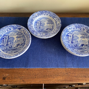 May include: A set of blue and white ceramic plates with a landscape design. The plates are displayed on a blue surface, presenting a classic, vintage style. The plates are round and feature a detailed, scenic illustration.