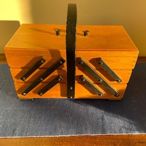 May include: A vintage wooden sewing box with a black handle and hinges. The box is made of light-coloured wood and has a tiered design with black metal accents. The box is sitting on a blue surface.