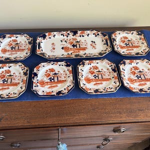 May include: A collection of vintage ceramic plates and a serving platter. The plates and platter have an orange and blue Asian-inspired design on a white background. The octagonal plates and rectangular platter are arranged on a blue cloth.