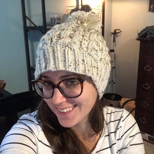 May include: A white knit beanie with a pom pom on top. The beanie has a cable knit pattern and is made of a soft, warm material.