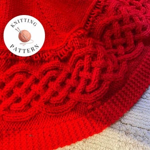 May include: A close-up of a red knitted blanket with a cable knit pattern. The blanket is folded in half and the edge of the blanket is visible. The blanket is made with a thick yarn and has a soft texture.