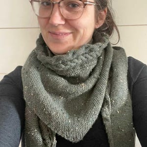 May include: A green knitted shawl with a speckled texture. The shawl is folded and draped around a person's neck.