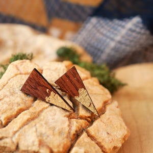 May include: A pair of wooden triangle earrings with a dark brown finish and gold leaf accents. The earrings are displayed on a textured rock surface, showcasing their geometric design and natural materials. These earrings are a unique accessory.