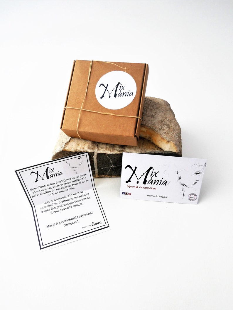 May include: A small brown box tied with twine and a white circle sticker with the text "Mix Mania" in black. The box is sitting on a brown rock. A white card with the text "Mix Mania bijoux & accessoires" in black is on a white surface. The card also has a small black and white illustration of a woman wearing earrings.