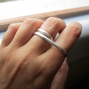 Puede incluir: Un anillo de plata con un diseño único y fluido. El anillo se envuelve alrededor de dos dedos, creando un aspecto moderno y artístico. El metal pulido refleja la luz, resaltando las curvas suaves y el estilo minimalista del anillo.