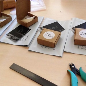 May include: Four small brown cardboard boxes with white labels, each with a different design. The boxes are on a white surface with business cards next to them. The business cards have a black and white photo of a woman and the text "Grazie!" and "Original".