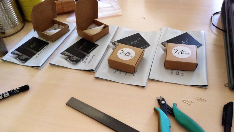 May include: Four small brown cardboard boxes with white labels, each with a different design. The boxes are on a white surface with business cards next to them. The business cards have a black and white photo of a woman and the text "Grazie!" and "Original".