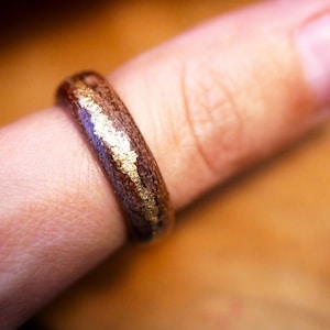 Puede incluir: Un anillo de madera con una base marrón oscuro y una incrustación dorada. El anillo se lleva en un dedo, mostrando el contraste entre la madera y el acento metálico. El diseño del anillo es simple pero elegante.
