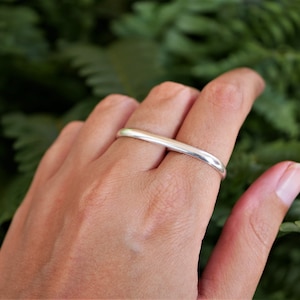 May include: A silver ring with a bar design worn on a finger. The ring is smooth and shiny, reflecting light. The background is a blurred green foliage.
