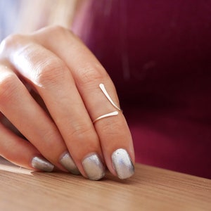 May include: A silver ring with a simple, minimalist design. The ring is made of two thin, curved pieces of metal that intersect in the middle. The ring is worn on the middle finger of a hand with silver nail polish.