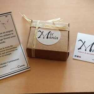 May include: A small brown box tied with twine and labeled "Mix Mania". The box is sitting on a light brown surface next to a white card with the text "Mix Mania" and instructions for cleaning silver and copper jewelry. A business card with the text "Mix Mania bijoux & accessoires" is also on the surface.