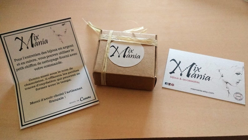 May include: A small brown box tied with twine and labeled "Mix Mania". The box is sitting on a light brown surface next to a white card with the text "Mix Mania" and instructions for cleaning silver and copper jewelry. A business card with the text "Mix Mania bijoux & accessoires" is also on the surface.