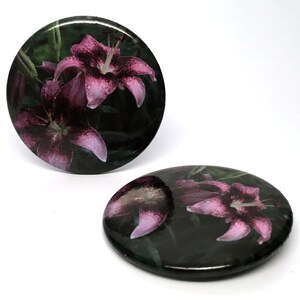 May include: Two round, black, glossy, decorative coasters featuring a purple lily flower design.