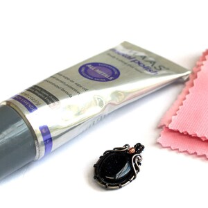 May include: A silver tube of jewelry polish with the text "IAS Jewel Polish" and "All Metals" on the label. A pink polishing cloth and a black gemstone pendant with silver wire wrapping are also pictured.