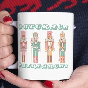 Puede incluir: Taza de cerámica blanca con cuatro figuras de cascanueces de dibujos animados en rosa, verde y beige. La taza tiene el texto "NUTCRACK PATRIARCHY" en una fuente verde menta. La taza es sostenida por una persona que lleva un suéter azul oscuro.