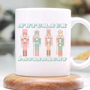 May include: White ceramic mug with the text "NUTCRACK the PATRIARCHY" and four illustrated nutcracker figures in pink, green, and gold. The mug sits on a round wooden coaster.