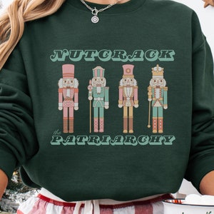 May include: Dark green sweatshirt featuring the text "NUTCRACK the PATRIARCHY" above a graphic of four nutcracker figures in pink, green, and gold. The sweatshirt has a crew neck.